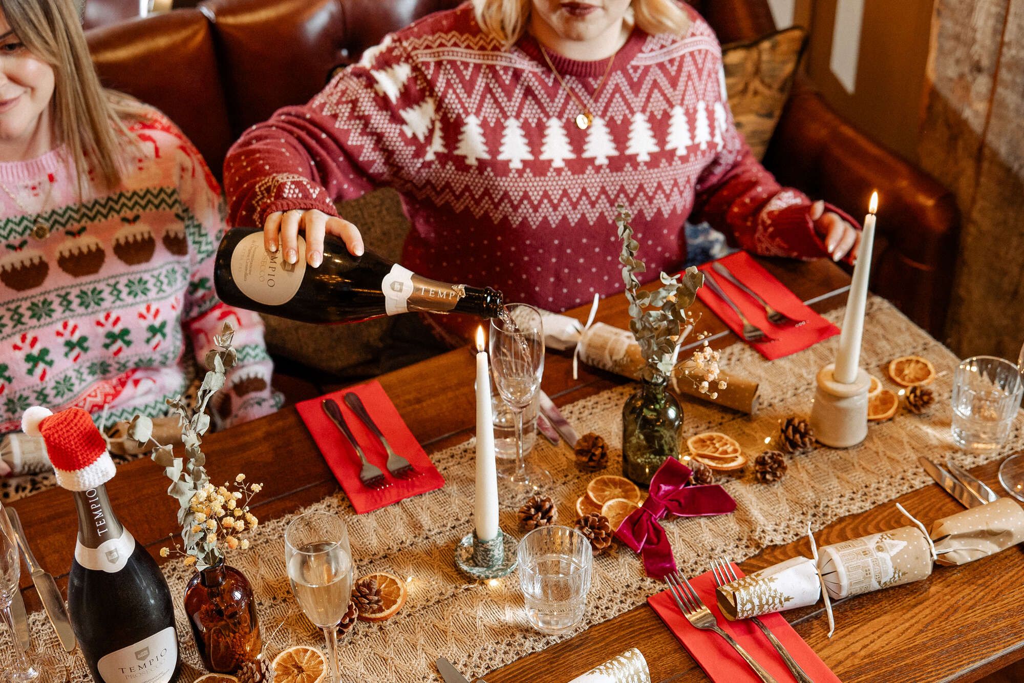 Christmas lunch table set with red napkins and crackers and someone pouring prosecco in a Christmas jumper
